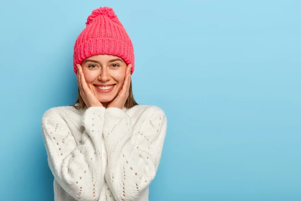 Smiling woman in a woollen jumper and pink beanie holding her face in her hands